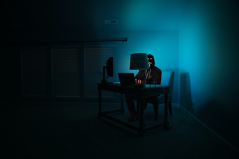 Eine Person mit Maske sitzt in einem verdunkelten Raum an einem Tisch vor einem Laptop. 